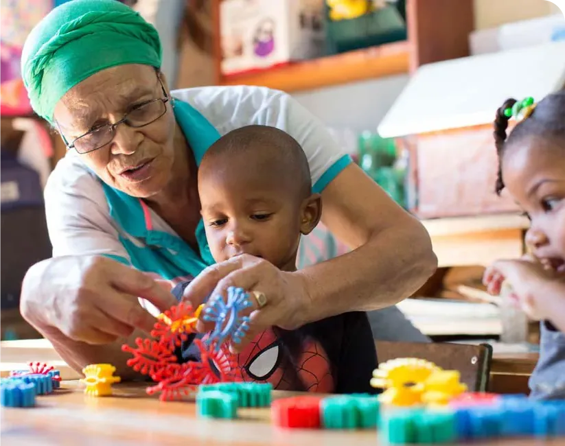 An older woman helps a young boy build with colourful plastic construction toys at a table.