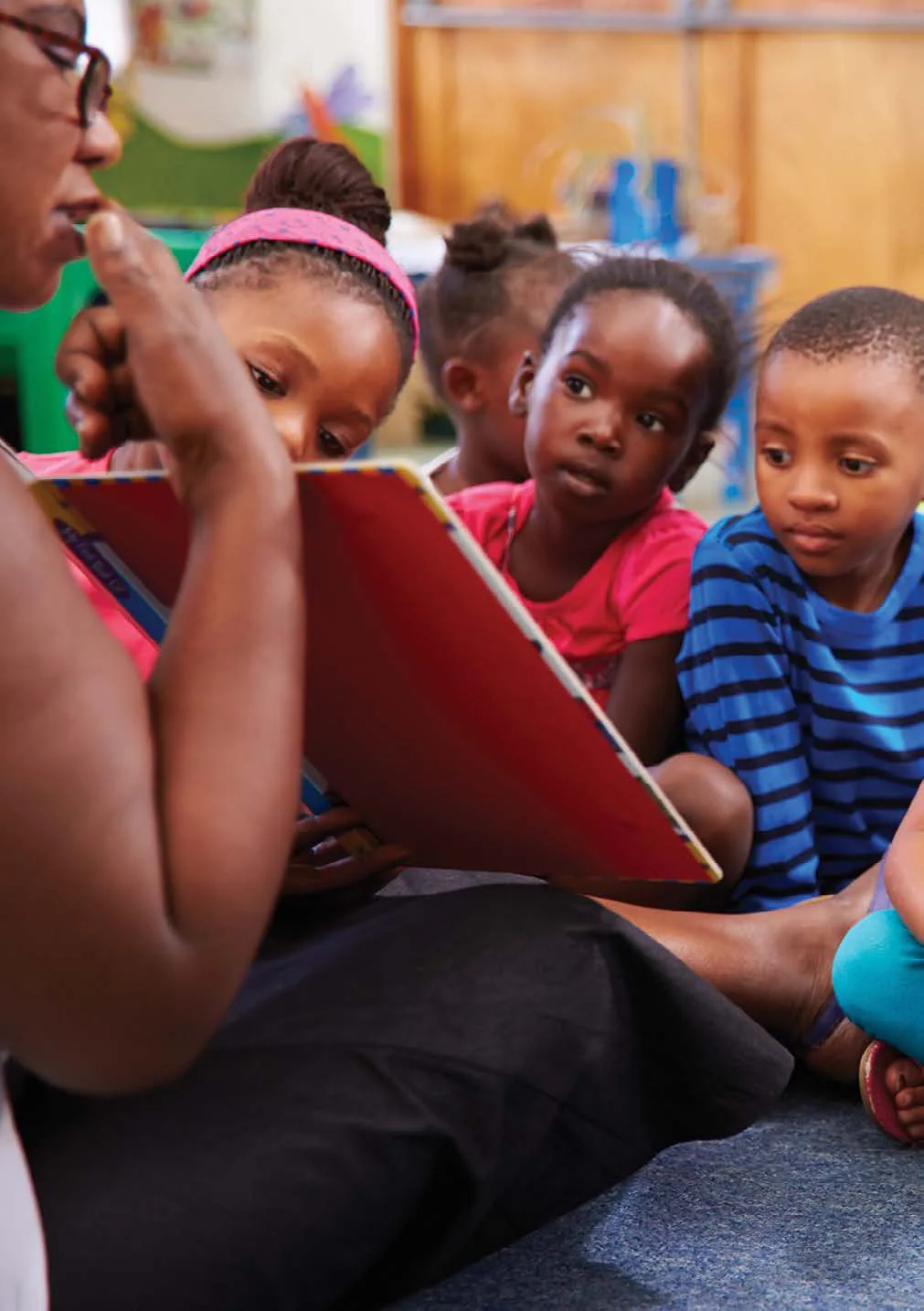 A woman reads a picture book aloud to a group of young children sitting close around her.