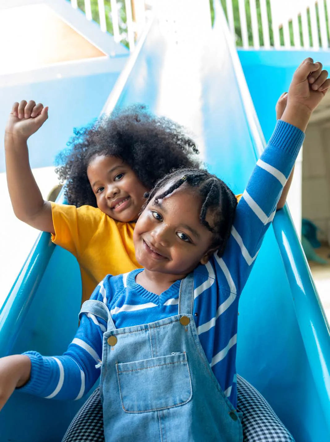 Two laughing young children slide down a blue playground slide with their arms raised.