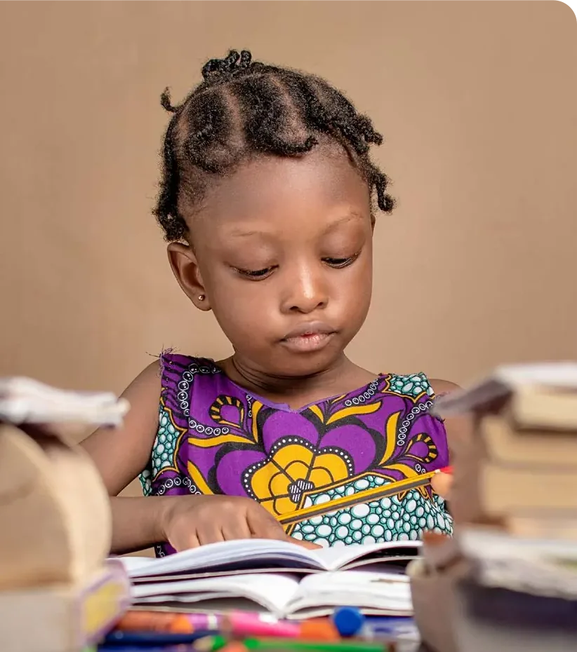 A young girl in a patterned dress concentrates on a book at a table.