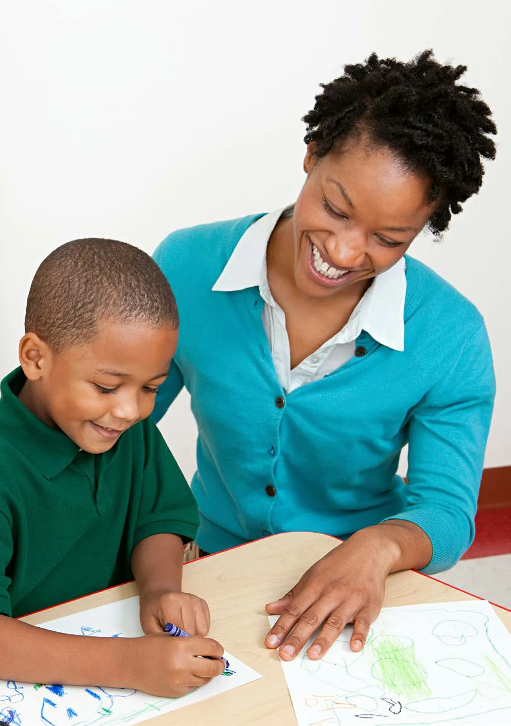 A smiling woman sits beside a young boy as he draws with crayons at a table.