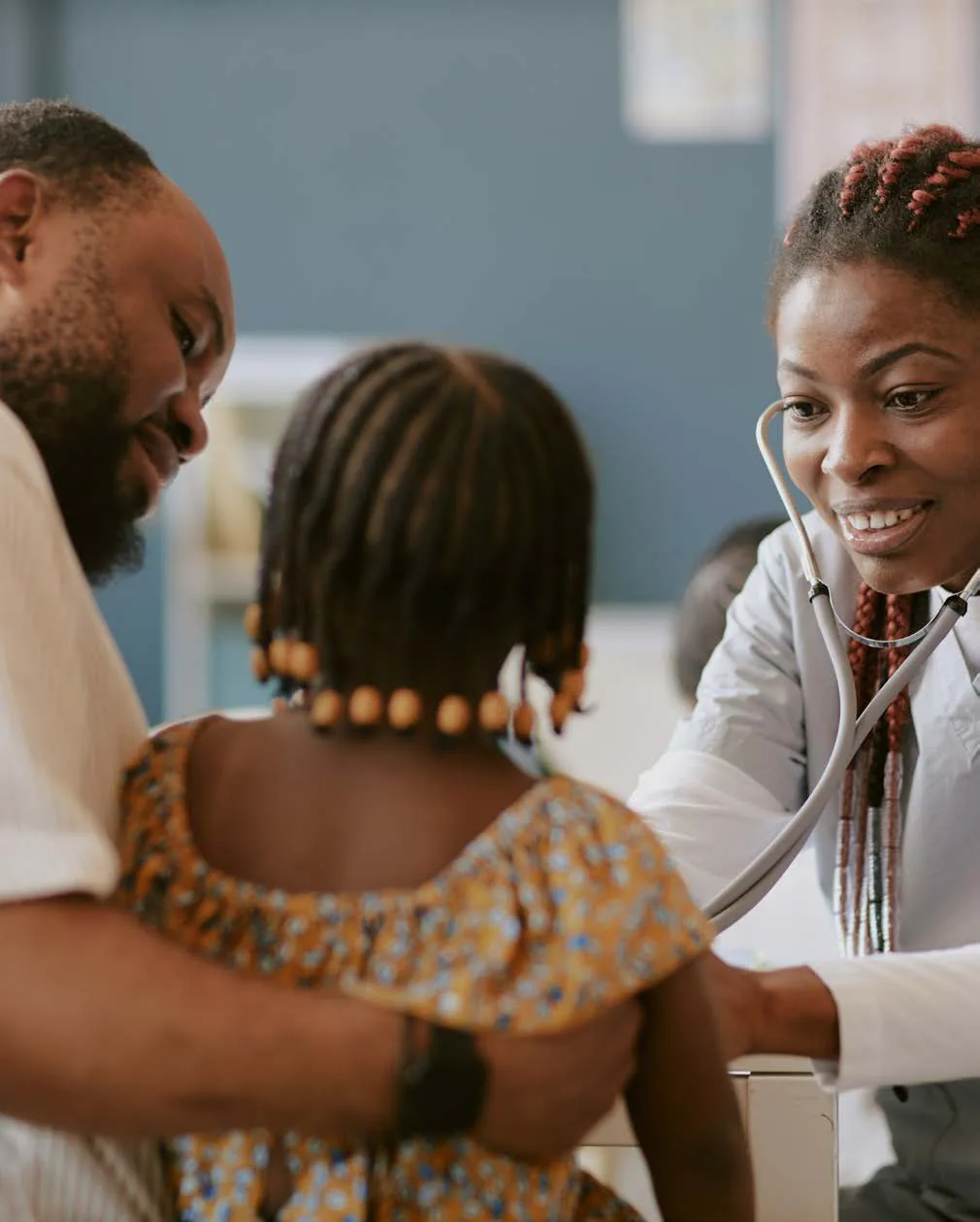A health worker with a stethoscope examines a young girl while her father holds her.