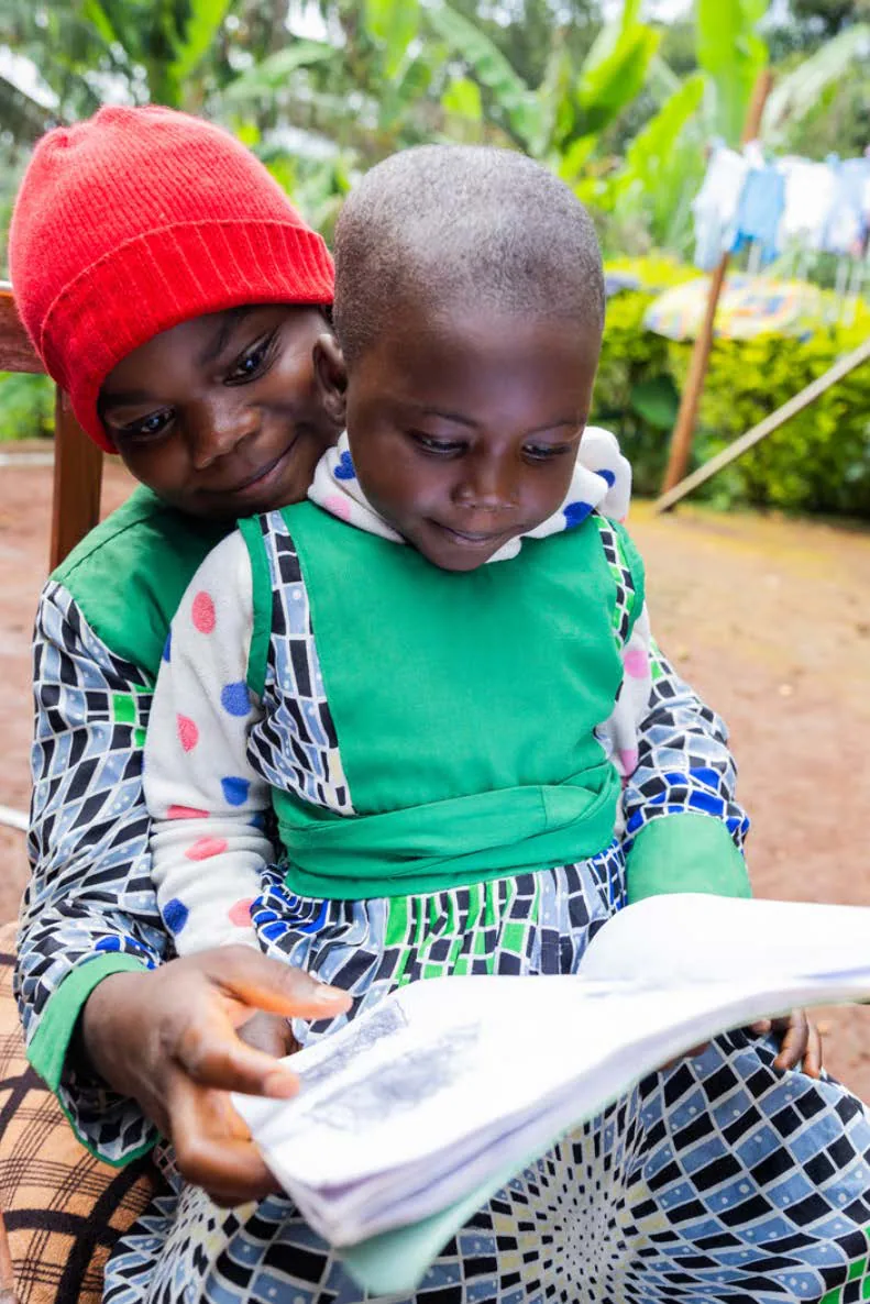 An older child in a red knitted hat reads a book with a younger child on her lap outdoors.