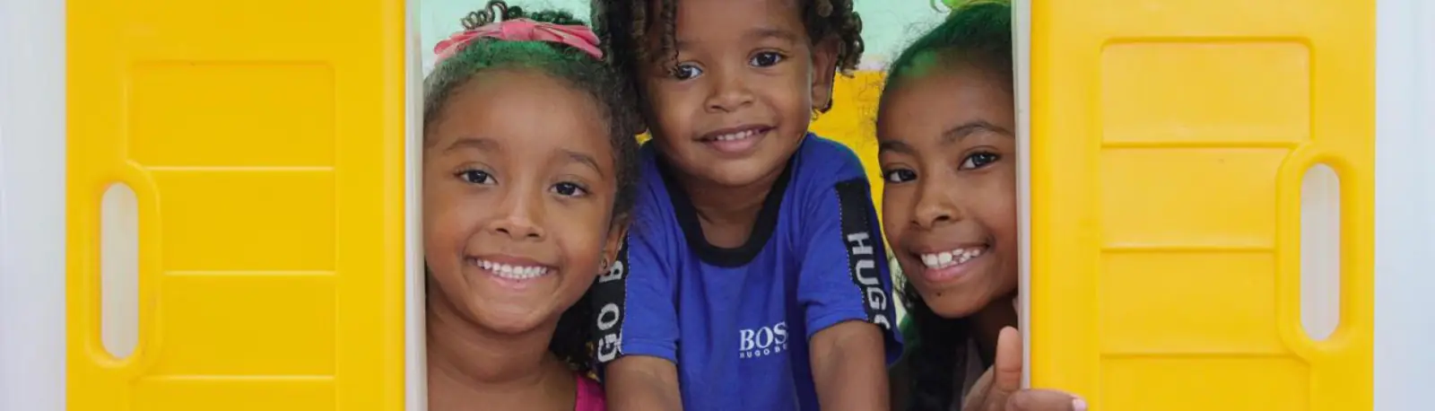 Three smiling children looking over a yellow wall.