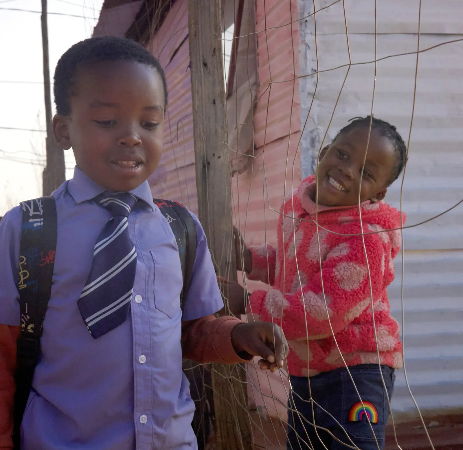 A boy in school uniform and tie and a smiling girl in a pink jacket beside a corrugated iron wall.