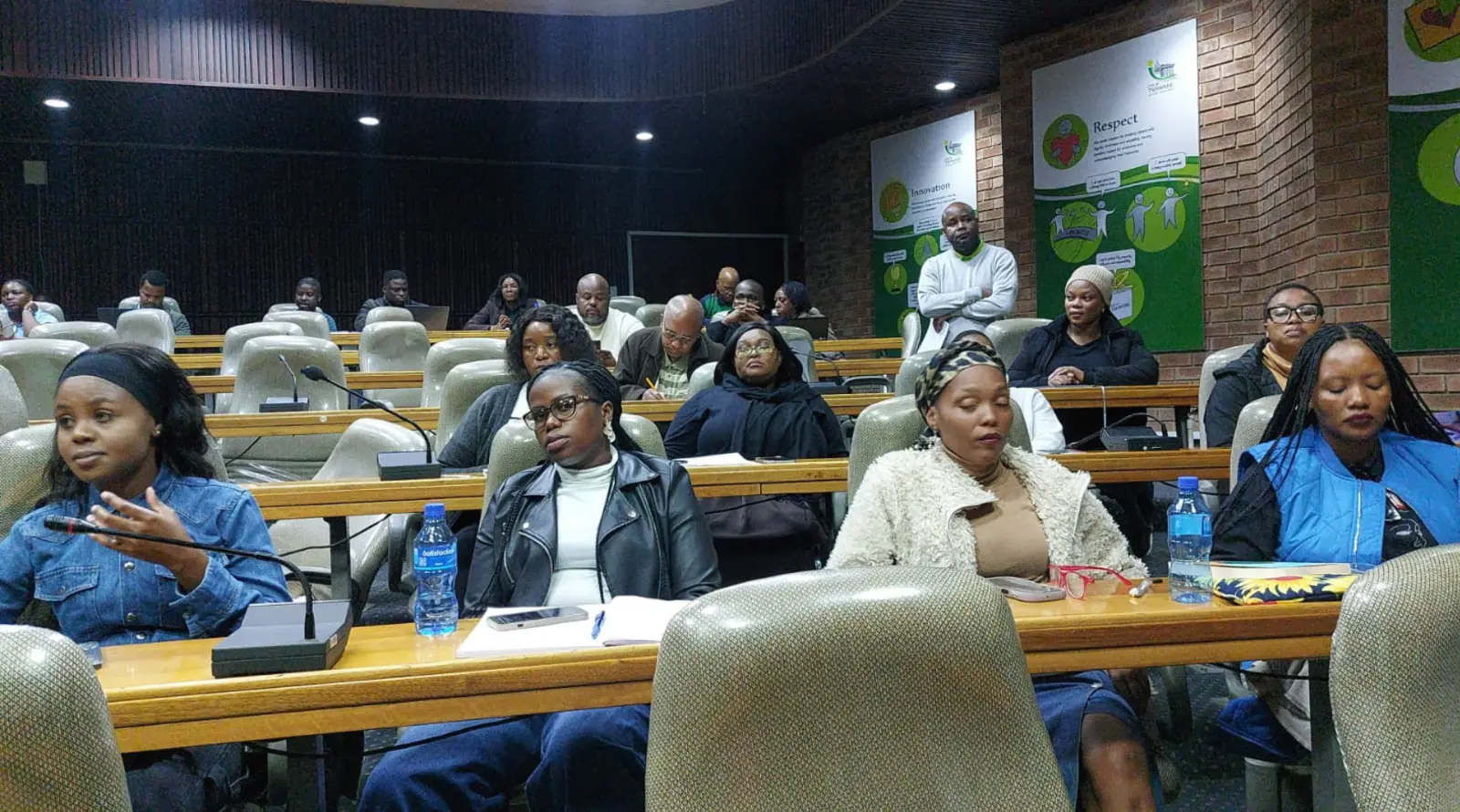 Workshop participants seated in rows of desks with microphones in a conference room.